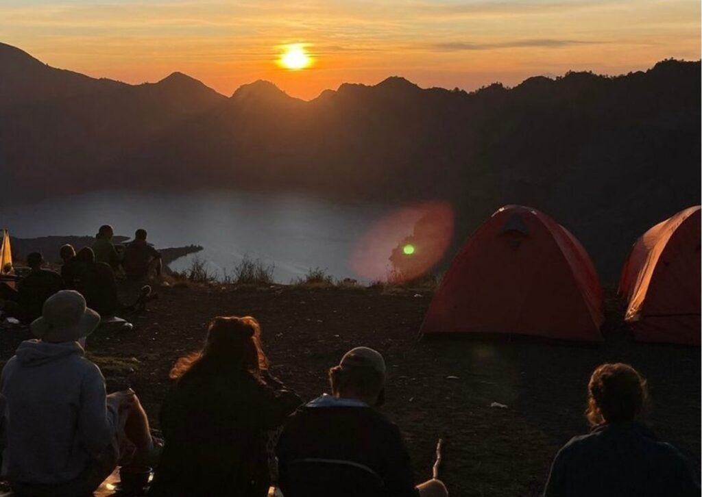 Golden sunset view from Sembalun campsite on Mount Rinjani with warm orange sky, mountain silhouettes, and tents overlooking the vast valley below